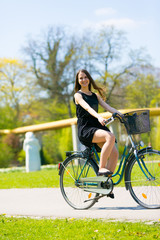Fototapeta na wymiar Portrait view of girl on bicycle wearing on black short dress. Young happy Woman riding along road on green spring outdoor Park. Sporty young girl riding a bicycle on a sunny morning, view from face