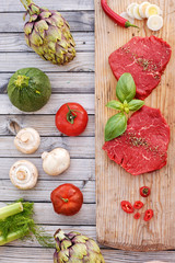 Top view of raw beef steaks on wooden board with fresh vegetables and champignons ready to be grilled. 