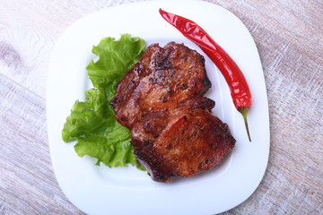 Steak of grilled meat and chili pepper with tomato, lettuce leaves on white plate.