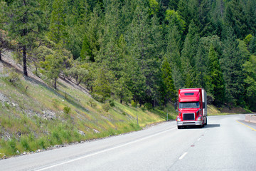 Modern red semi truck with trailer move cargo uphill on awesome green highway with forest trees on...