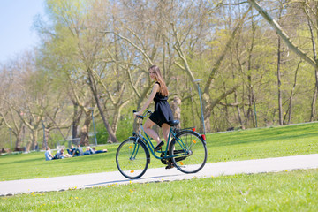 Portrait view of girl on bicycle wearing on black short dress. Young happy Woman riding along road on green spring  outdoor Park. Sporty young girl riding a bicycle on a sunny morning