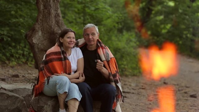 Elderly couple sitting covered with a blanket on the log, in the foreground a fire is burning.