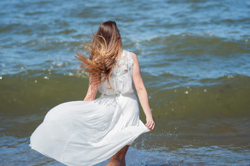 Young charming brunette woman on sea coast. Beautiful girl in a white summer dress. Runs towards the sea, back view