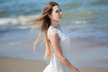 Fototapeta na wymiar Young charming brunette woman on sea coast. Beautiful girl in a white summer dress. On the background of sea waves