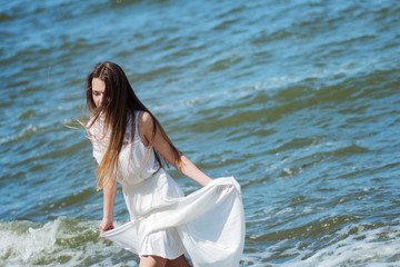 Young charming brunette woman on sea coast. Beautiful girl in a white summer dress. On the background of sea waves