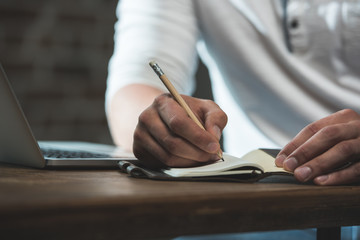 Cropped shot of man writing in notebook during work while sitting at the table with laptop