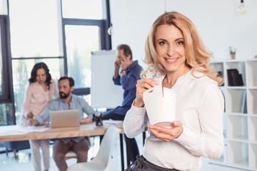 Smiling blonde businesswoman eating asian food while colleagues working behind