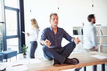 Middle aged businessman sitting on table and meditating in lotus position while colleagues working...