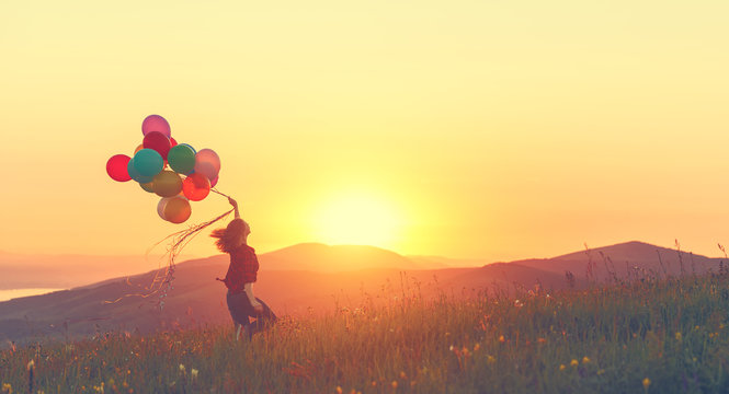 Happy cheerful girl with balloons running across meadow at sunset in summer