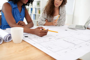 Multi-ethic group of engineers gathered together in open plan office and working on blueprint, wooden desk on foreground