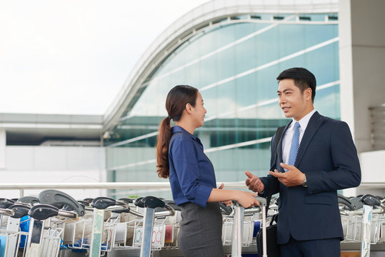 Portrait of modern Asian business partners, man and woman, talking in airport, smiling and gesturing actively
