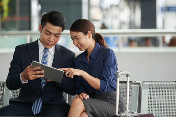Portrait of two Asian business people, man and woman,  using digital tablet while waiting in airport departure zone travelling on business trip