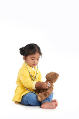Full length portrait of serious toddler sitting in lotus position and listening to heartbeat of her teddy bear with help of stethoscope, isolated on white background