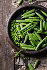 Young organic green pea pods and peas in terracotta tray over old dark wooden planks with sackcloth textile background. Top view with space. Harvest, healthy eating.