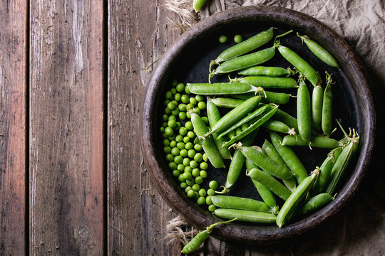 Young organic green pea pods and peas in terracotta tray over old dark wooden planks with sackcloth textile background. Top view with space. Harvest, healthy eating.