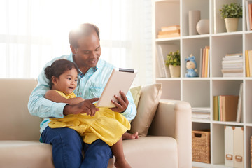 Family portrait of cute toddler sitting on laps of her middle-aged father and watching cartoons on digital tablet, interior of cozy living room on background