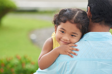 Cheerful little girl with deep black eyes looking away while hugging her dad, green lawn of public...