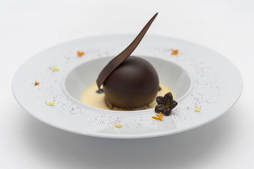 Luxurious dessert with chocolate globe and vanilla sauce, served on a biscuit, decorated with chocolate flower, flower petals and forest fruits, served in a white plate, light background, isolated