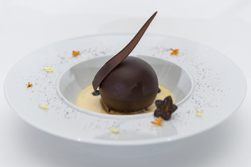 Luxurious dessert with chocolate globe and vanilla sauce, served on a biscuit, decorated with chocolate flower, flower petals and forest fruits, served in a white plate, light background, isolated