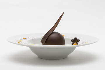 Luxurious dessert with chocolate globe and vanilla sauce, served on a biscuit, decorated with chocolate flower, flower petals and forest fruits, served in a white plate, light background, isolated