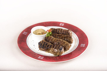 Mici, traditional romanian meat rolls, served with mustard, decorated with green and red leafs, placed on a white and red plate, light background