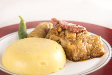Sarmale, a traditional Romanian stuffed cabbage dish, served with polenta, bacon and hot chilli, placed on a white and red plate, light background
