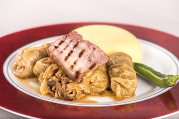 Sarmale, a traditional Romanian stuffed cabbage dish, served with polenta, bacon and hot chilli, placed on a white and red plate, light background