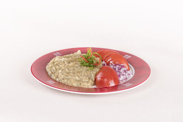 Eggplant salad, served with red onion and sliced tomatoes, decorated with green and red leafs, placed on a white and red plate, light background