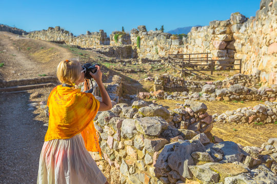 Traveler woman photographer takes shot of Archaeological Site of Ancient Mycenae, Peloponnese, Greece. Caucasian beauty blonde female photographing a ruins of defence stone walls. Europe Heritage Site