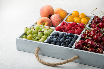Assorted summer fruits and berries in a light wooden box with cells standing on light concrete background. Blueberries, raspberries, gooseberries, peaches, cherries, alycha