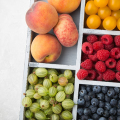 Assorted summer fruits and berries in a light wooden box with cells standing on light concrete background. Blueberries, raspberries, gooseberries, peaches, alycha. Top view