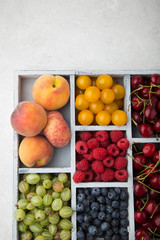 Assorted summer fruits and berries in a light wooden box with cells standing on light concrete background. Blueberries, raspberries, gooseberries, peaches, cherries. Top view