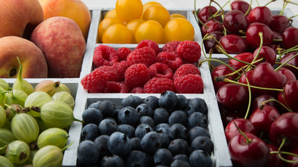 Assorted summer fruits and berries in a light wooden box with cells standing on light concrete background. Blueberries, raspberries, gooseberries, peaches, cherries, alycha