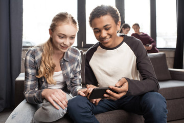Happy teenage couple using smartphone on sofa while friends standing behind, teenagers having fun concept