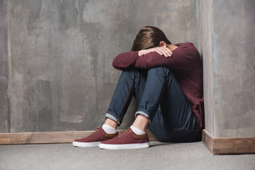 Depressed teenage boy sitting on floor with head in hands - obrazy, fototapety, plakaty