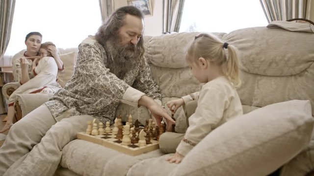Bearded man playing chess with little son on couch in the living room while loving mother hugging little daughter sitting on her knees