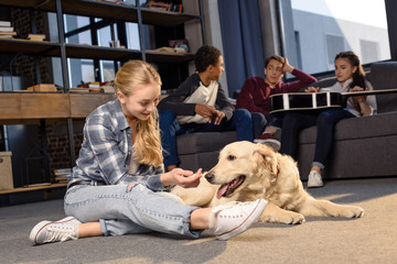 Smiling teenage girl having fun with golden retriever dog while friends sitting on sofa