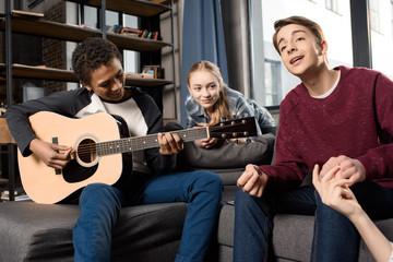 african american boy playing acustic guitar while his friends listening and singing at home, teenagers playing guitar concept