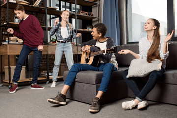 african american boy playing acustic guitar while his friends dancing behind at home, teenagers playing guitar concept