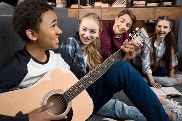 african american boy playing acustic guitar and singing while his friends listening at home,...