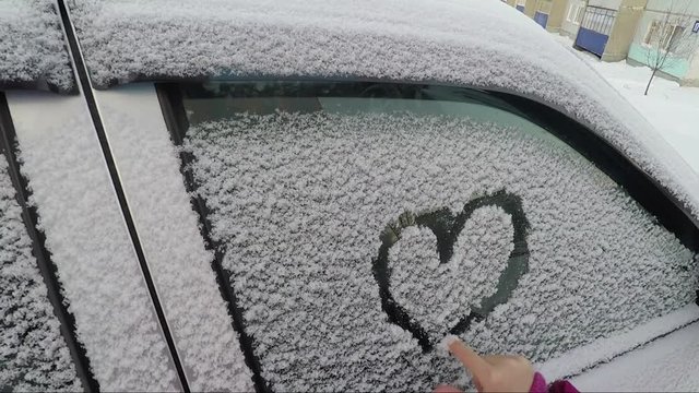 A child's hand paints a heart on a winter car glass