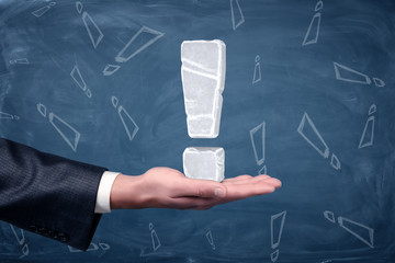 An exclamation mark made of white ice placed on businessman's open palm on chalkboard background.
