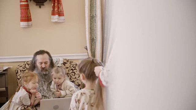 Caring mother doing fishtail braid for little daughter; little boy and girl watching something on laptop with dad. Family wearing traditional Russian clothing at home