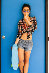 Fototapeta premium Young sexy stylish hipster woman with skateboard look to the camera and make silence sign with finger near lips in front of blue wooden door. Swag urban teen girl. Outdoor lifestyle portrait.