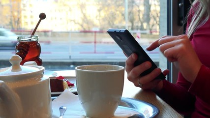 A woman sits at a table with meal in a cafe and works on a smartphone - closeup from the side, cars drive across a window in the background