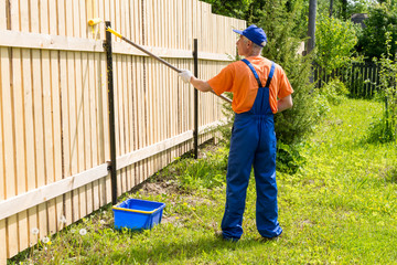 Full-length portrait of painter wearing blue dungarees, orange t-shirt, cap and gloves painting a wooden wall with paint roller.