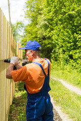 Close up portrait of handy carpenter fixes wooden fence with cordless electric screwdriver