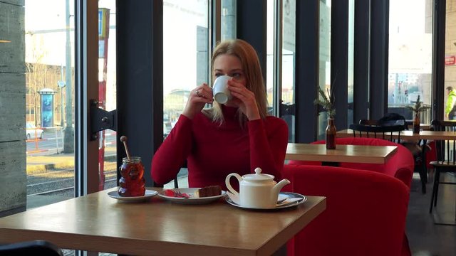 A young attractive woman sits at a table with meal in a cafe, drinks tea and looks out a window with a smile