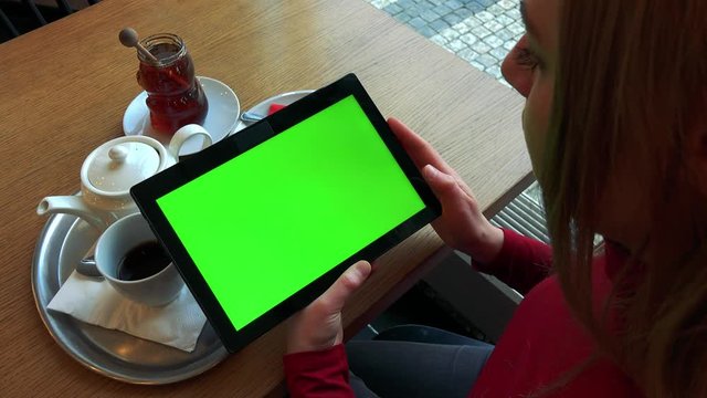 A blonde woman sits at a table with meal in a cafe and watches a tablet with a green screen - closeup from above