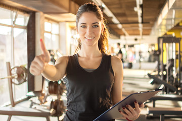 Portrait of personal trainer with clipboard showing thumb up at gym.